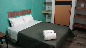 Una cama bien hecha con una colcha verde y almohadas blancas, una mesita de noche pequeña y un cabecero de madera.