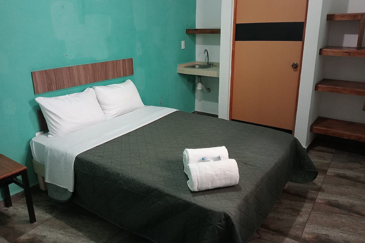 A neatly made bed with a green bedspread and white pillows, a small bedside table, and a wooden headboard.