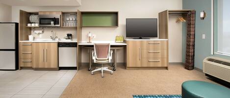 A compact hotel room with a kitchenette, a desk with a chair, a TV, a wardrobe, and a window with curtains.