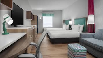 Een hotelkamer met een bureau, twee bedden, een grijze bank, een gestreept voetenbankje en een televisie.