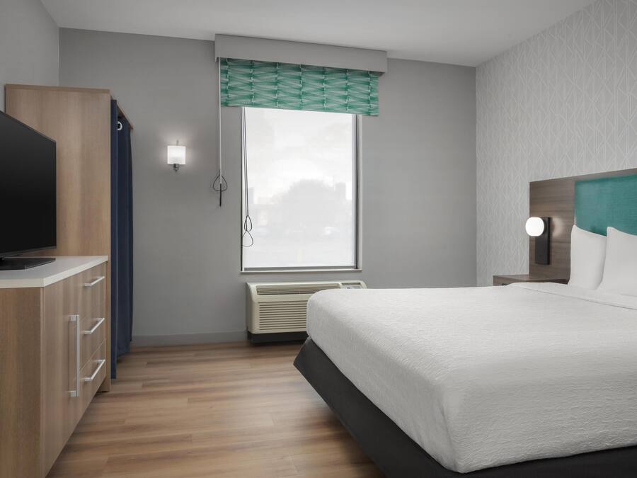 A modern hotel room with a bed, a TV, a wardrobe, a window with a view, and a wall-mounted light.