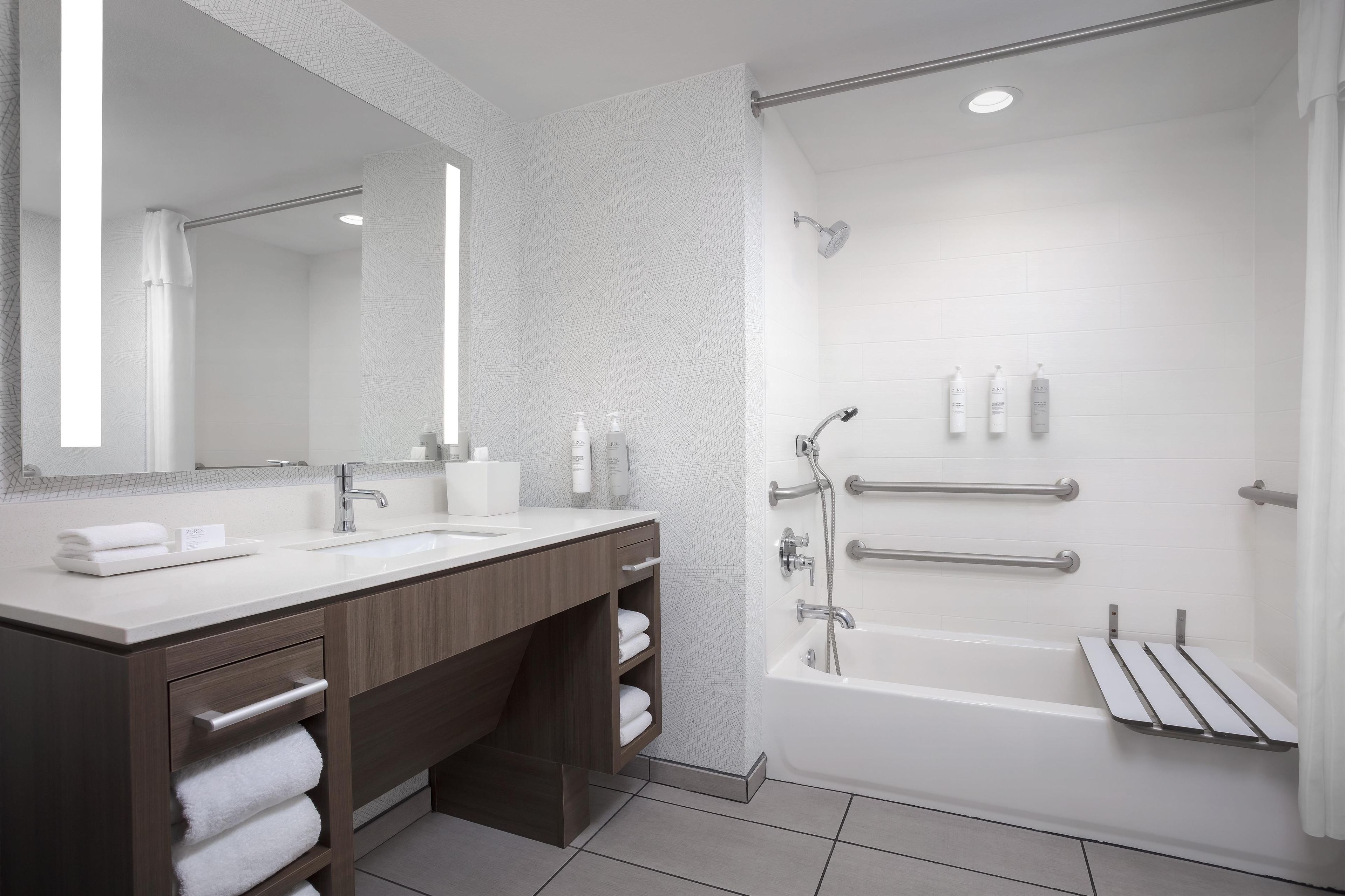 Une salle de bain moderne dotée d’un meuble-lavabo à deux vasques, d’un grand miroir, d’une douche équipée de barres d’appui et d’une baignoire.