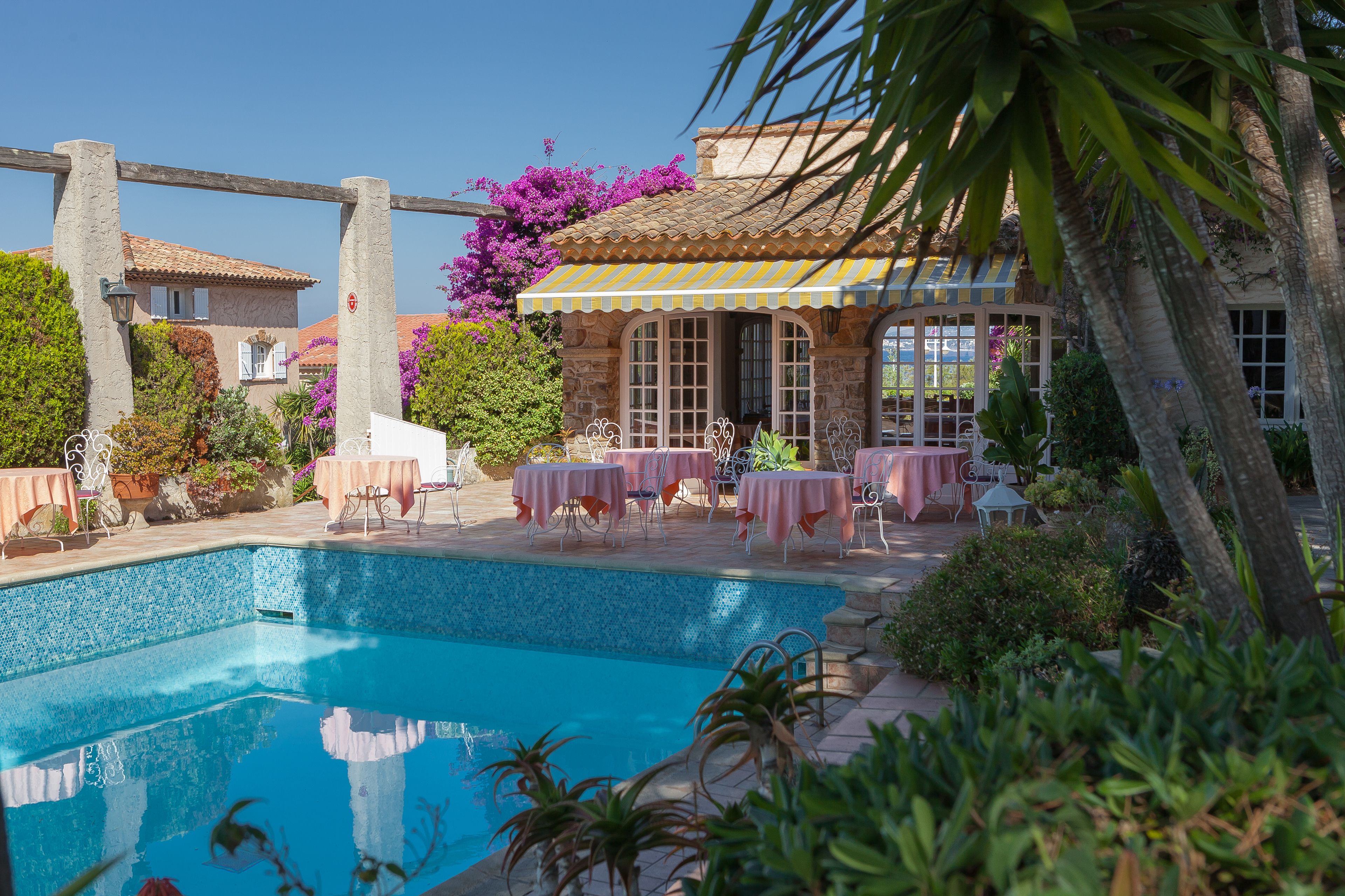 A swimming pool with tables and chairs, surrounded by lush greenery and a building with a tiled roof.