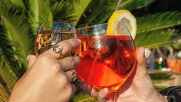 Two people holding glasses of a red beverage with a lemon slice, outdoors with greenery in the background.