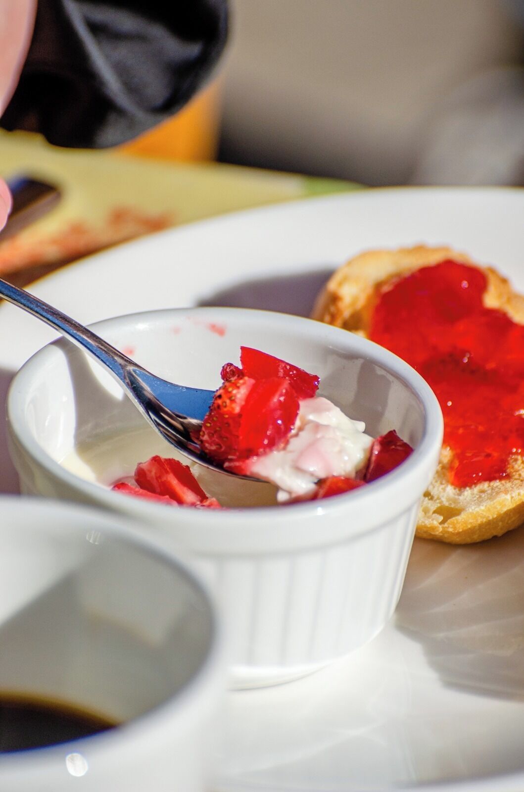 A dessert with strawberries and cream in a white bowl, a slice of toast with jam, and a spoon.