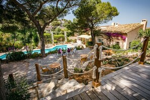 A wooden deck with a pool, surrounded by lush greenery and a rustic building with a tiled roof. - Hôtel Escapade (Le Pradet)