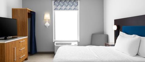 A hotel room with a bed, a TV on a wooden dresser, a window with a view, and a wall-mounted lamp.