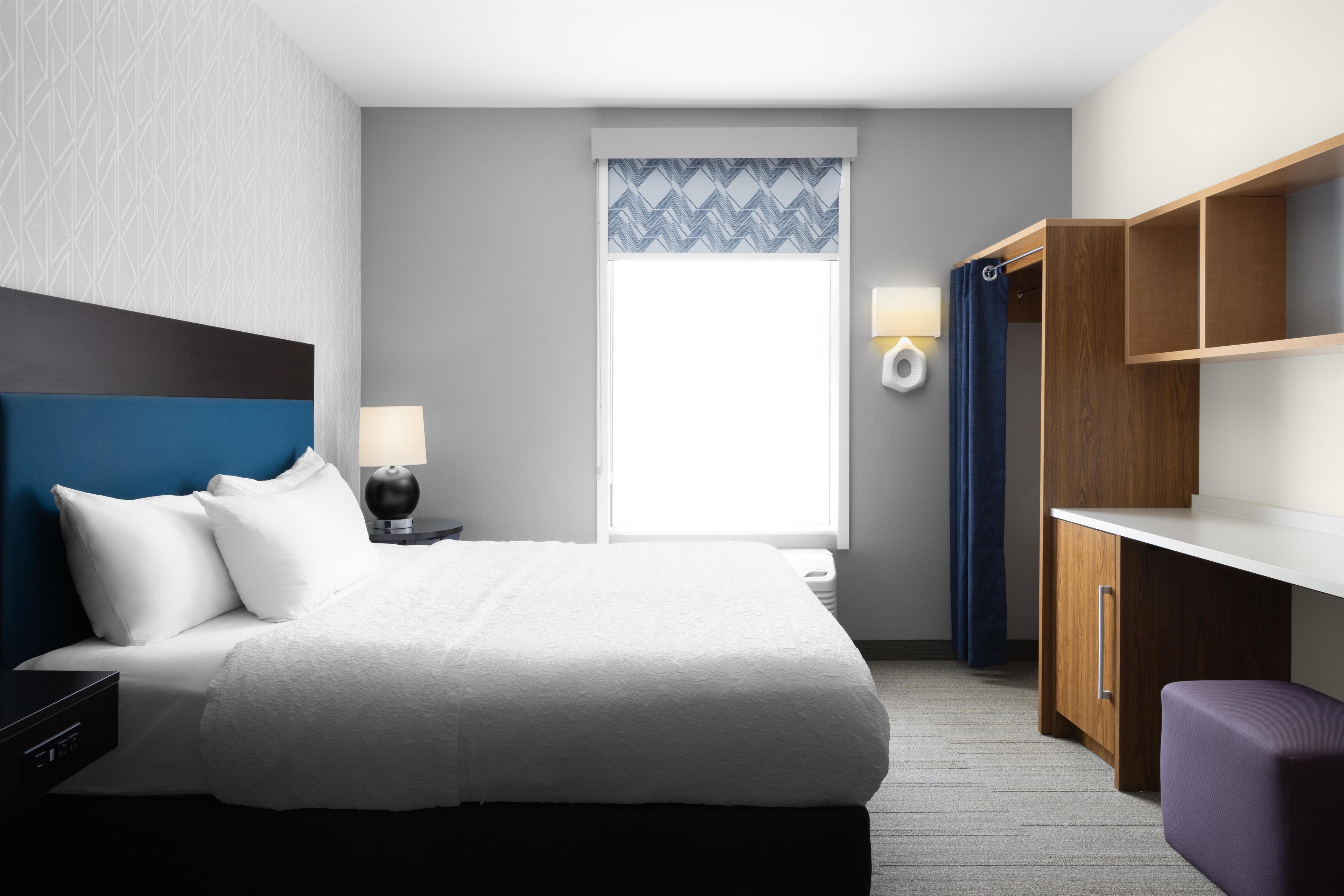 A hotel room with a bed, bedside table, lamp, wardrobe, and a window with a view.