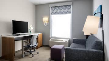A hotel room with a desk, chair, computer, lamp, couch, and a window with a view.