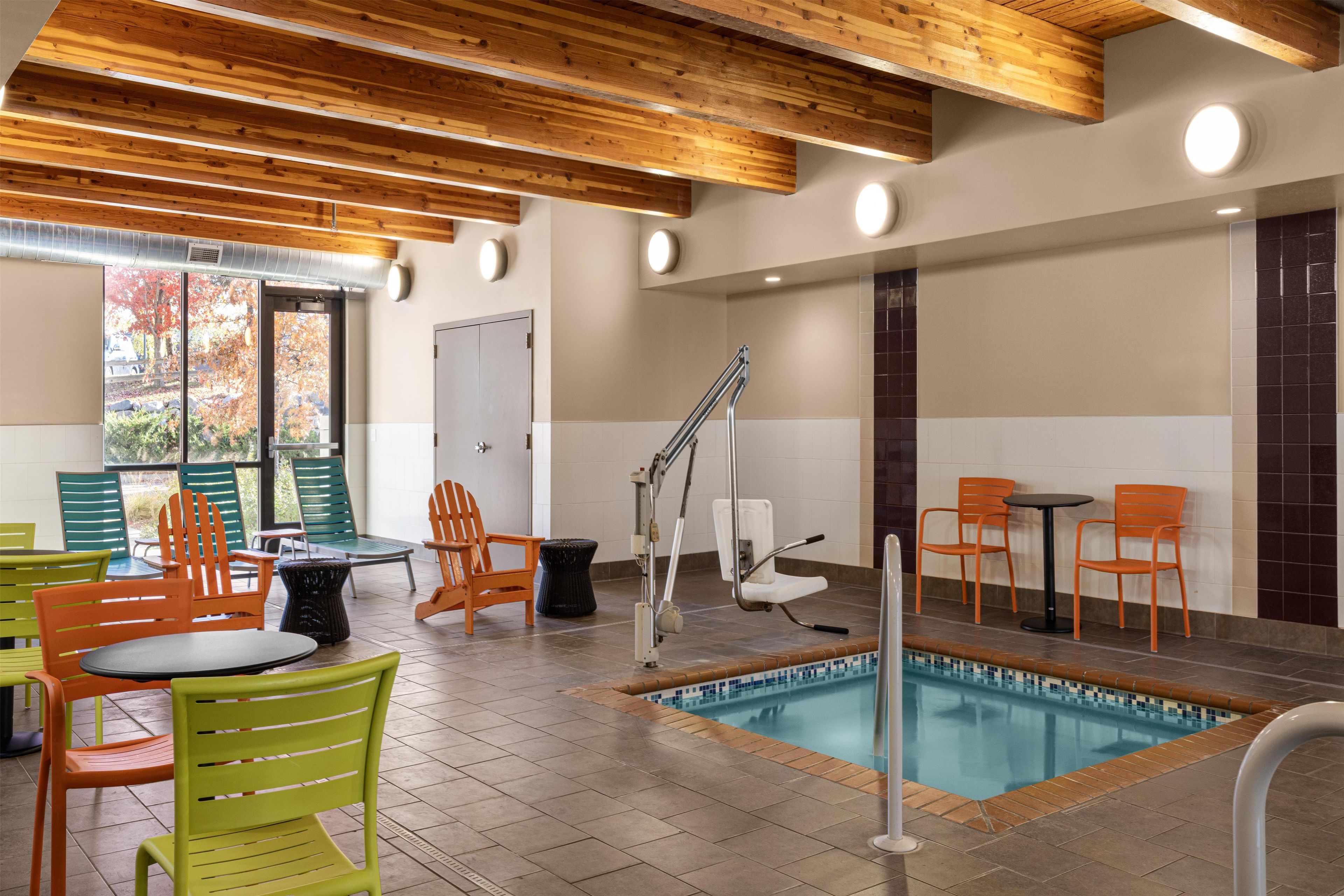 A pool area with chairs and tables, wooden ceiling beams, and a view of trees outside.