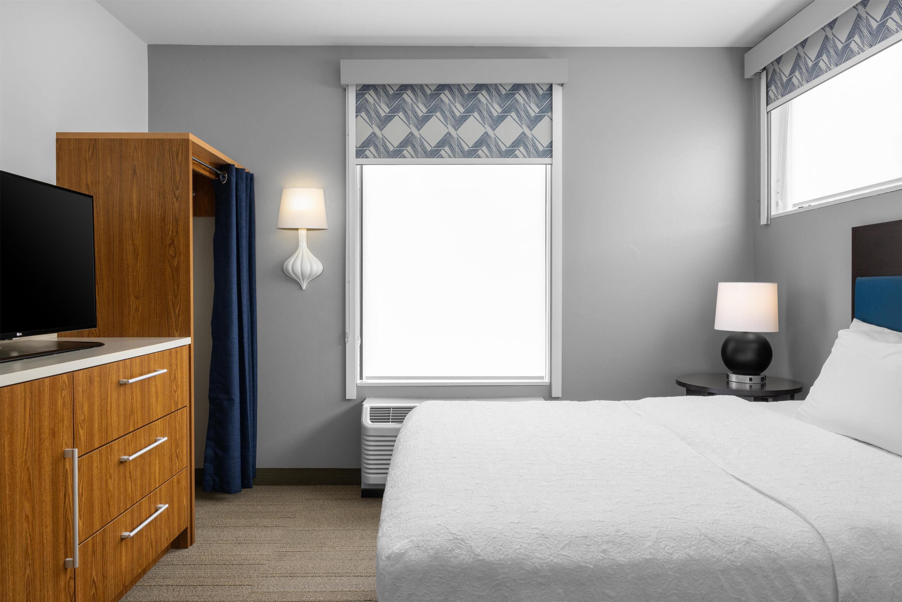 A hotel room with a bed, a TV on a wooden cabinet, a window with curtains, a bedside lamp, and a wall-mounted heater.