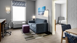 A modern hotel room with a gray patterned wall, a large window with a view, a gray sofa, a purple ottoman, a wooden table, and a floor lamp. - Home2 Suites by Hilton Minneapolis-Eden Prairie (Minnetonka)
