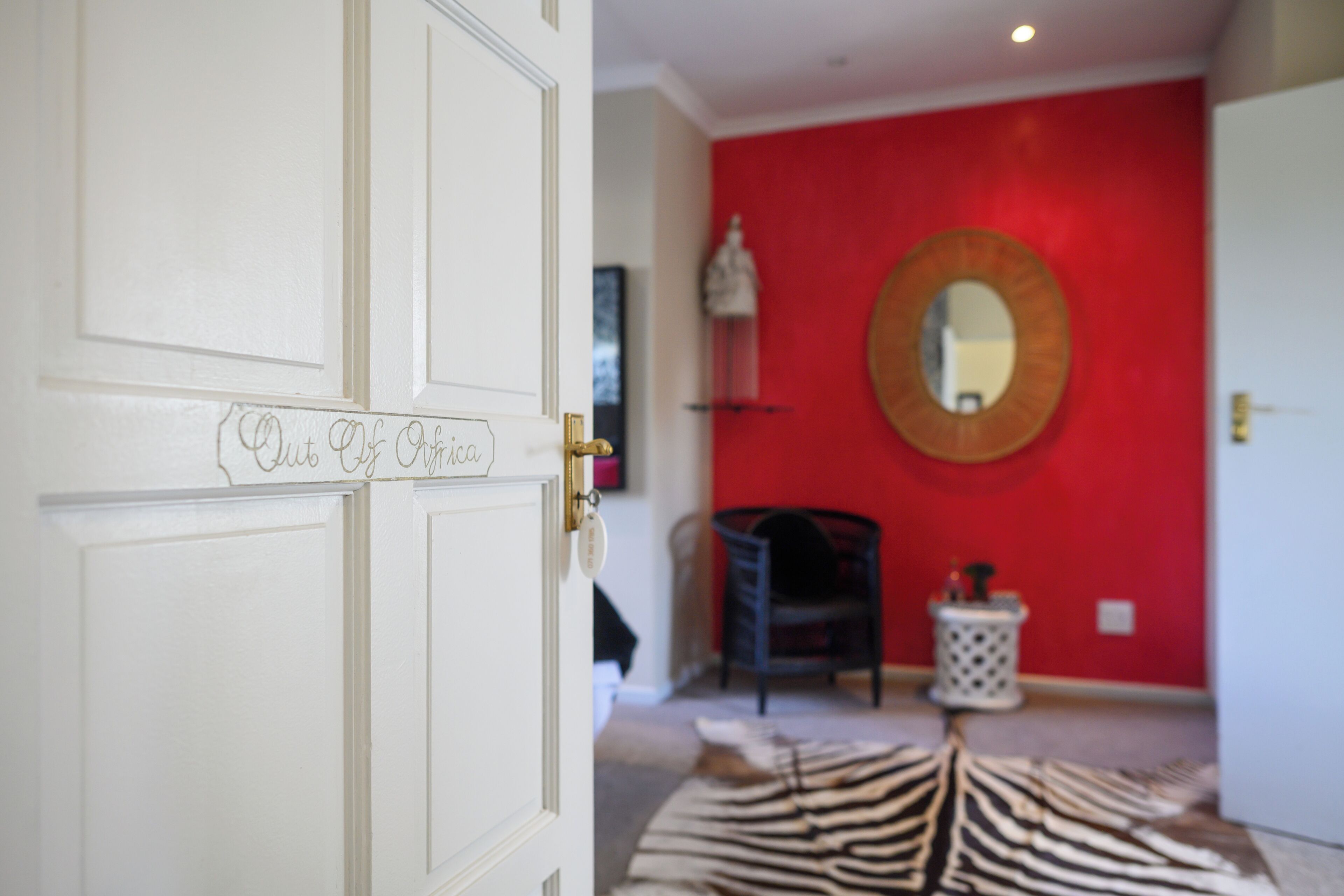 Una habitación con una puerta blanca, una pared roja, un espejo redondo, un felpudo con estampado de cebra y un cubo de basura blanco pequeño.