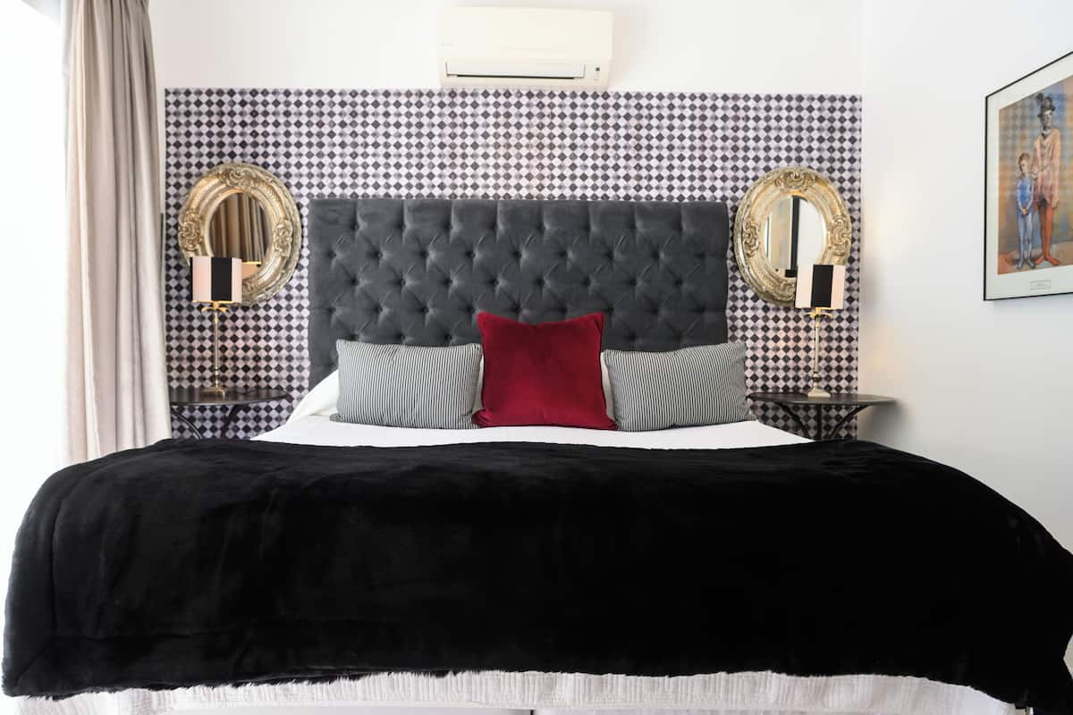 Une chambre à coucher avec une tête de lit capitonnée, deux miroirs fixés au mur, un coussin décoratif rouge et un plaid noir.