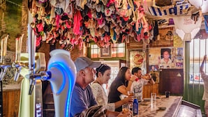 A bar with a variety of clothing items hanging from the ceiling, a counter with a few people, and a sign that reads "Merry Christmas." - Paternoster Hotel (Paternoster)
