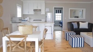 Een moderne keuken met witte kastjes en een centraal eiland, een eethoek met houten stoelen, en een zithoek met een zwarte bank en gestreepte poufs.
