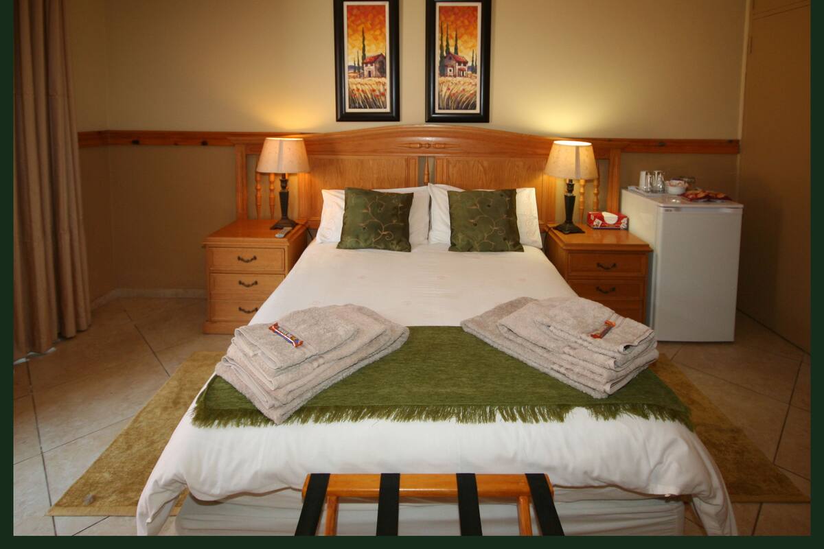 A hotel room with a bed, two nightstands, a lamp, a refrigerator, and two paintings on the wall.