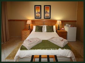Habitación de hotel con cama, dos mesitas de noche, una lámpara, un refrigerador y dos cuadros en la pared.