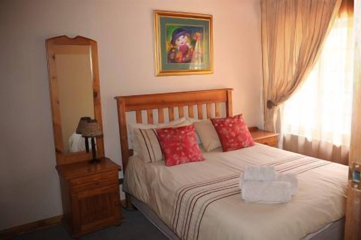 A bedroom with a wooden headboard, a bed with striped bedding, two red pillows, a nightstand with a lamp, a mirror, and a painting on the wall.