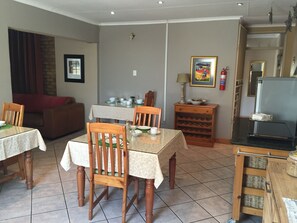 Comedor con una mesa dispuesta para cuatro personas, rodeada de sillas de madera. Se observa un mueble bajo con copas de vino y una lámpara sobre una mesita auxiliar. En el fondo, un sofá y un cuadro enmarcado son visibles.