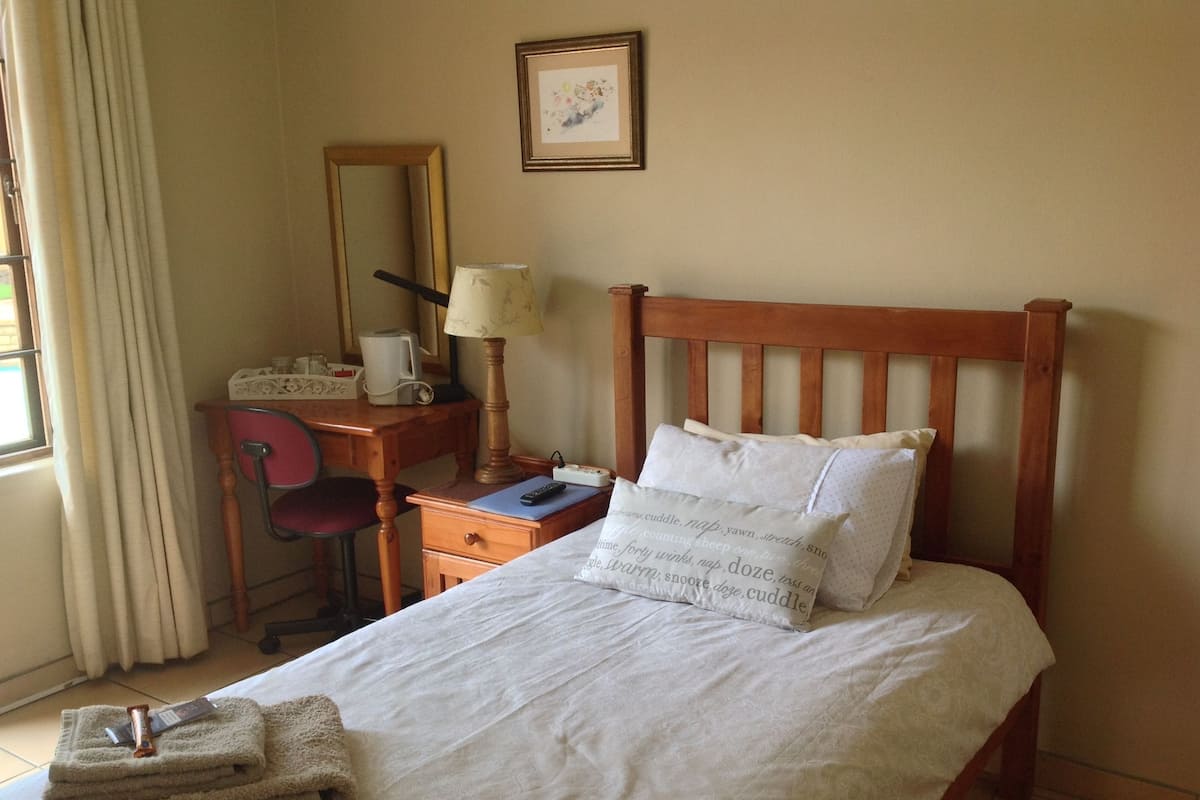A neatly made bed with pillows, a wooden headboard, a bedside table with a lamp, a chair, and a desk with a mirror and a framed picture on the wall.