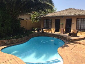 Una piscina con borde curvo, rodeada por un patio de ladrillo. Hay un banco y una mesa pequeña cerca de la piscina.