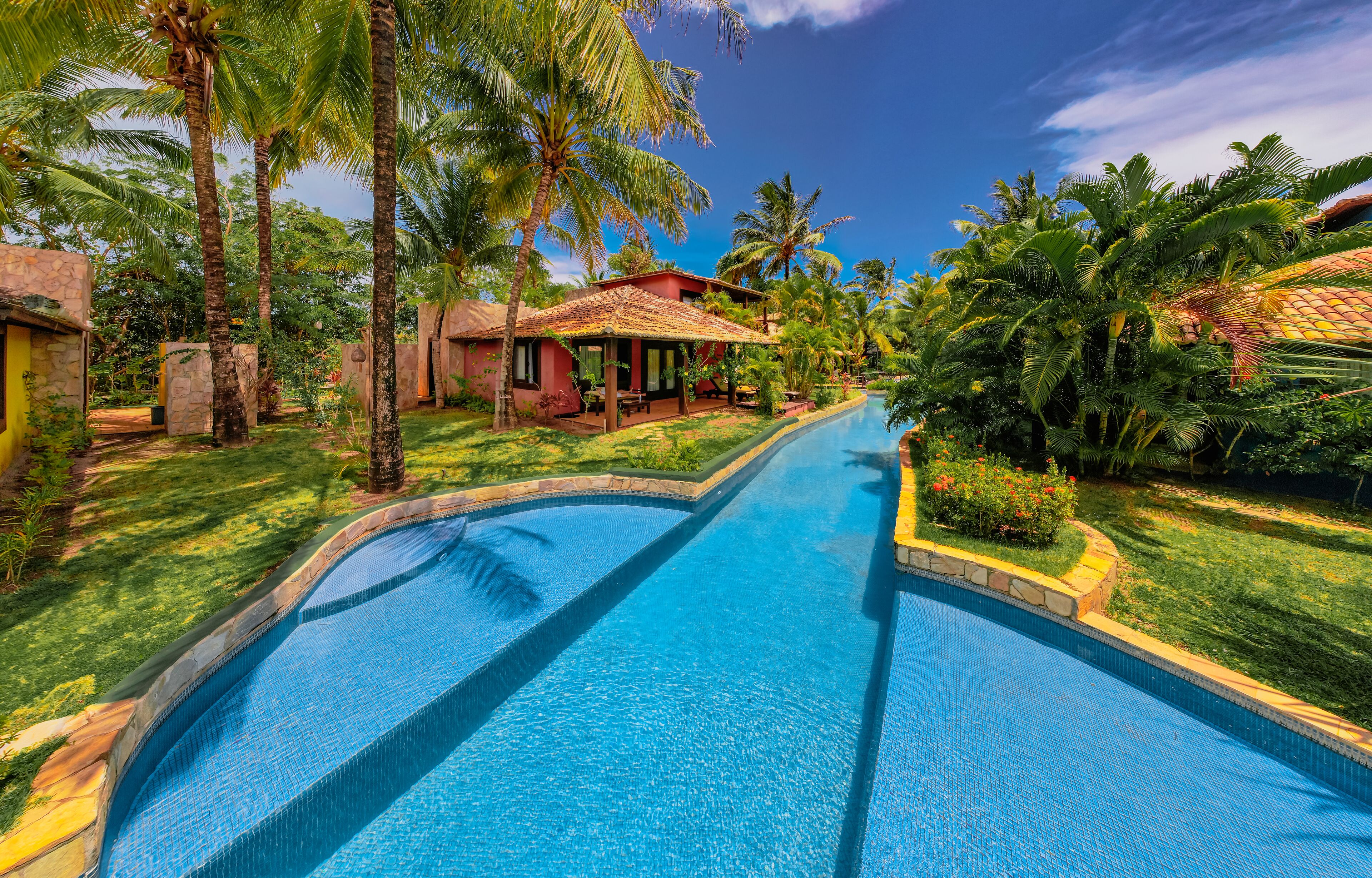 Una piscina rodeada de exuberante vegetación y palmeras, con una casa al fondo.