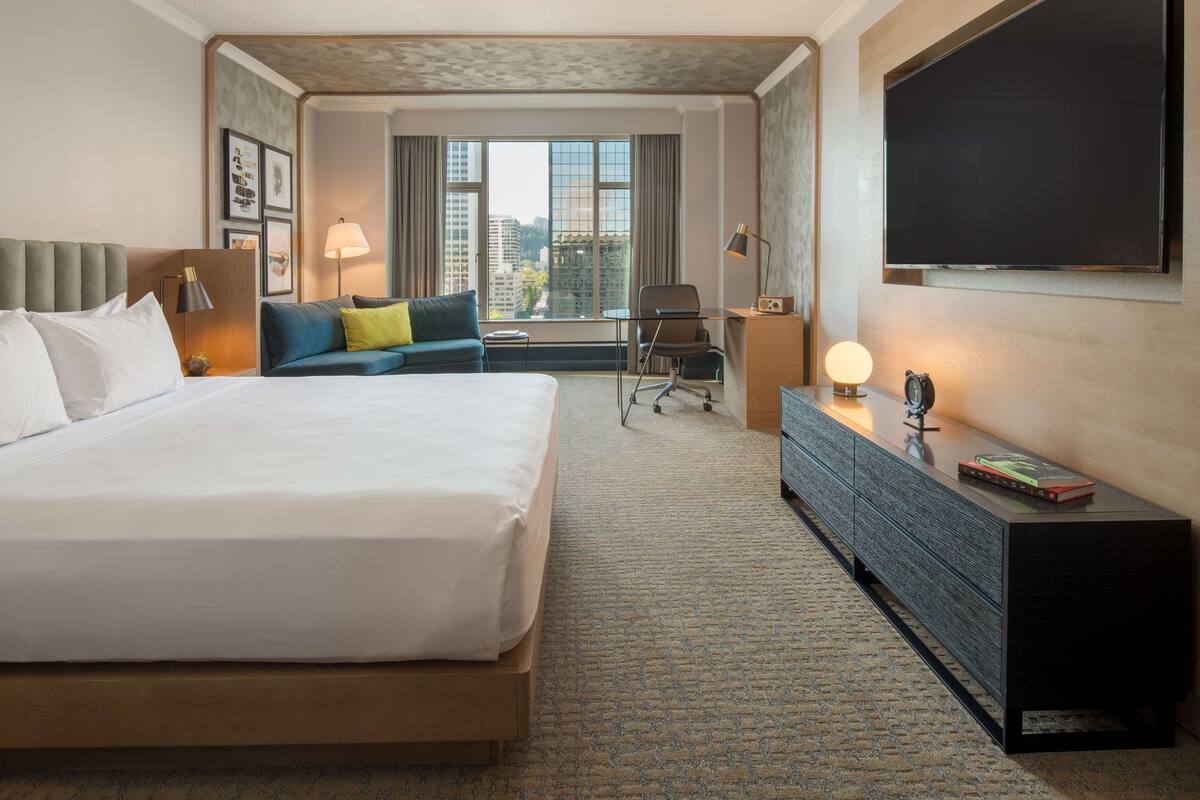 A hotel room with a large bed, a desk, a chair, a TV, and a window with a city view.