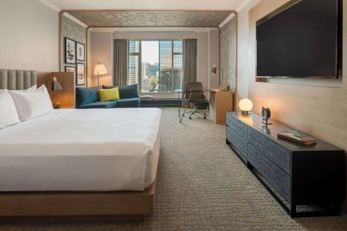 A hotel room with a large bed, a sofa, a desk, a TV, and a window with a city view.