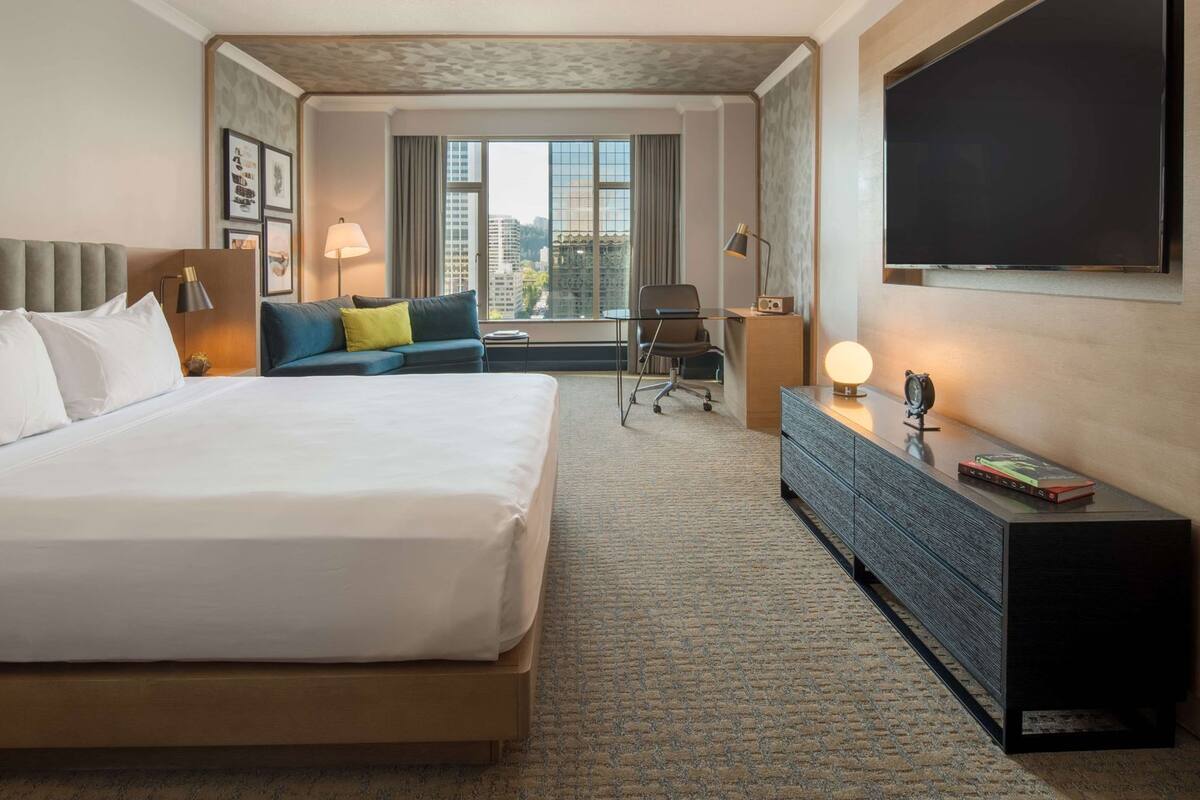 A hotel room with a large bed, a sofa, a desk, a TV, and a window with a city view.