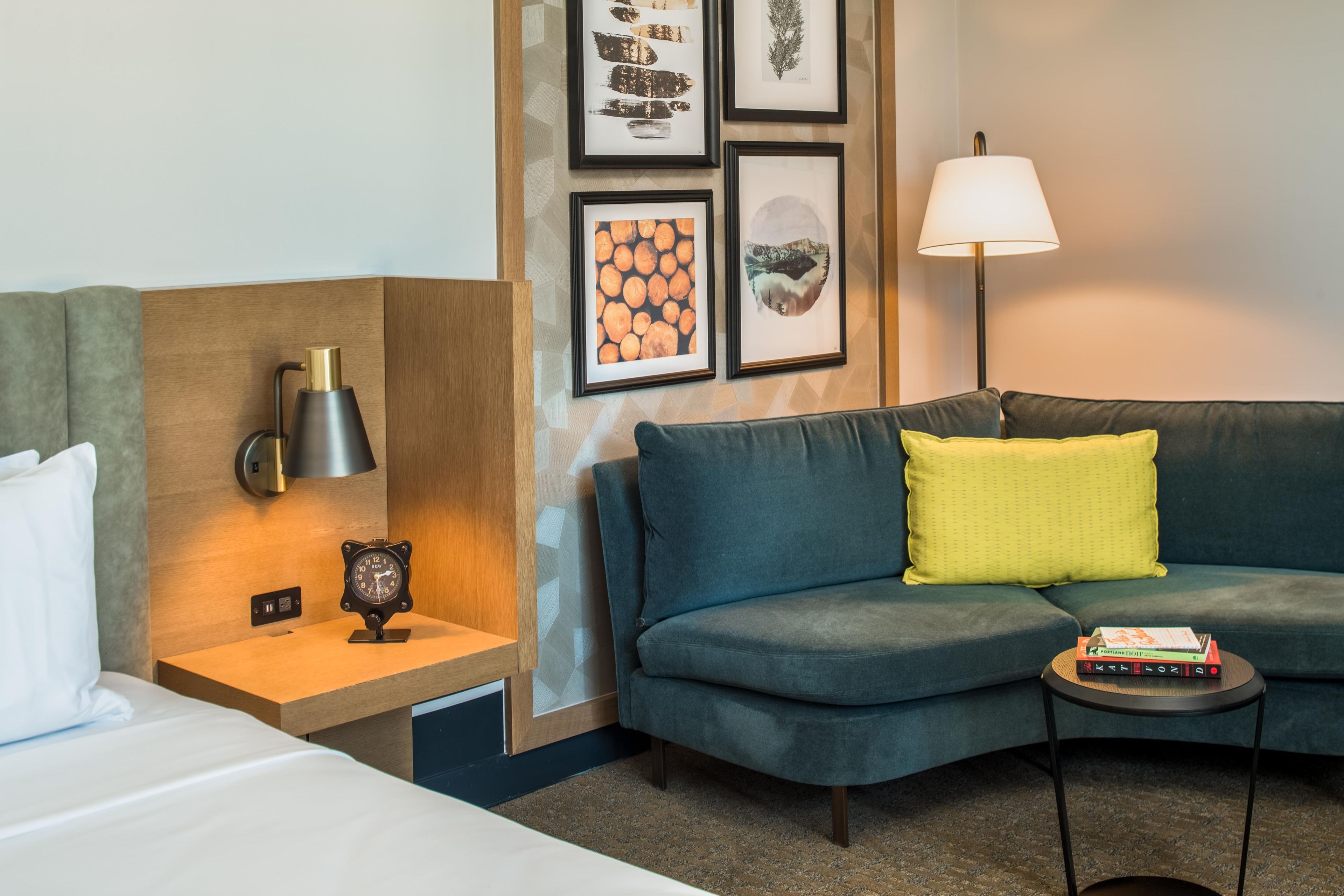 A hotel room with a bed, a nightstand with a lamp, a sofa with cushions, a small side table, and a wall with framed pictures.