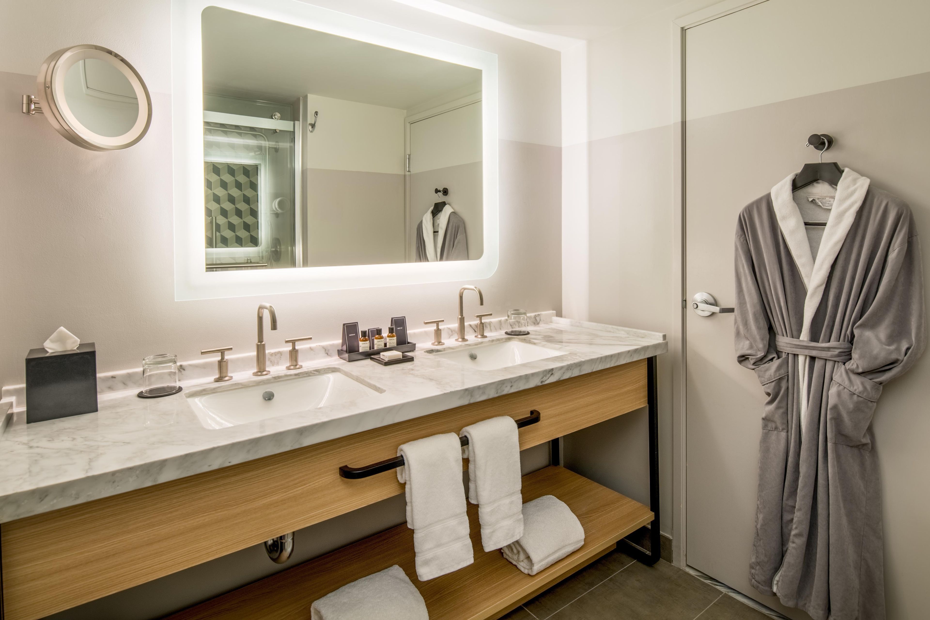 A modern bathroom with a large mirror, two sinks, a robe hanging, and a towel rack.