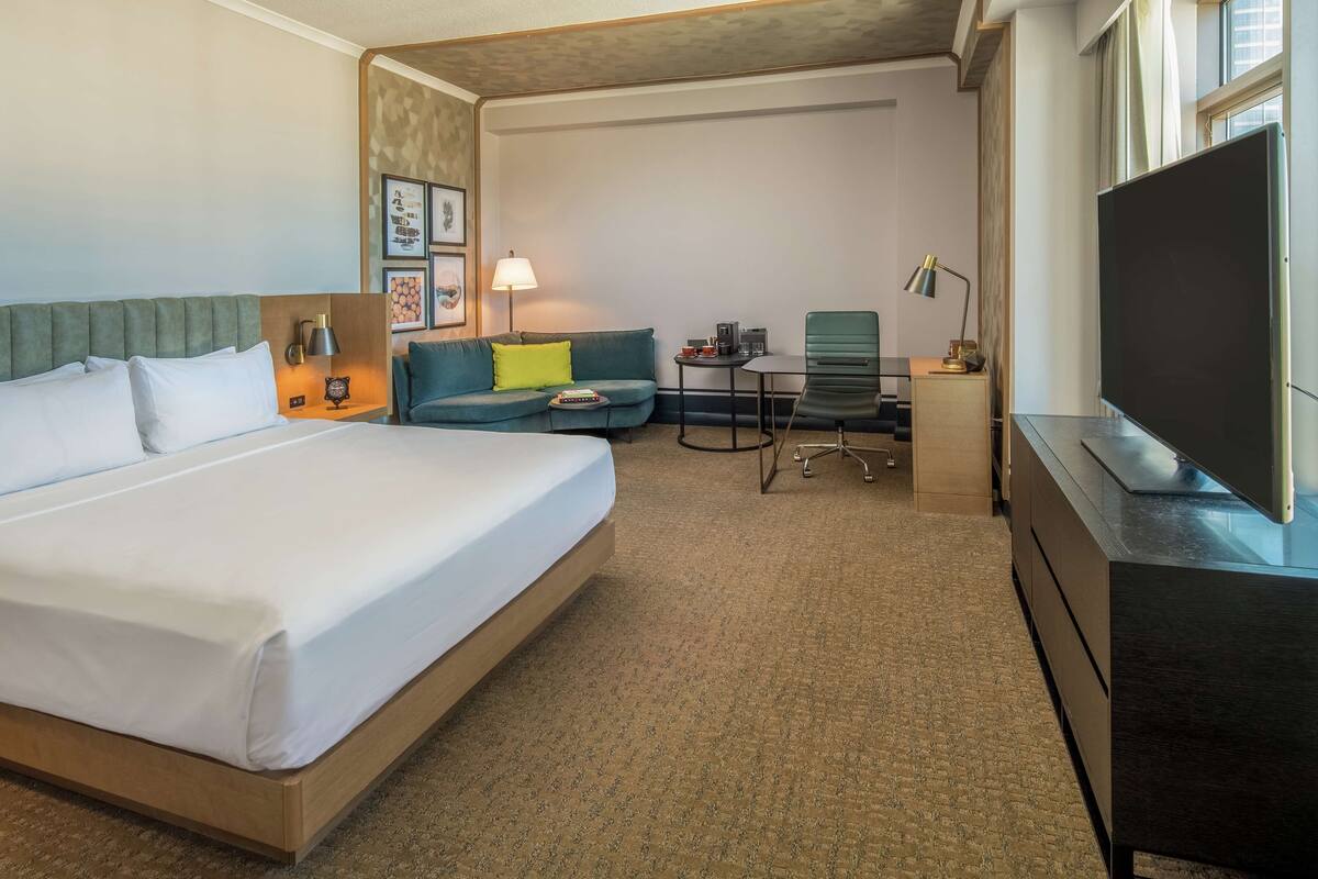 A hotel room with a large bed, a sofa, a small table with a lamp, a chair, and a TV.