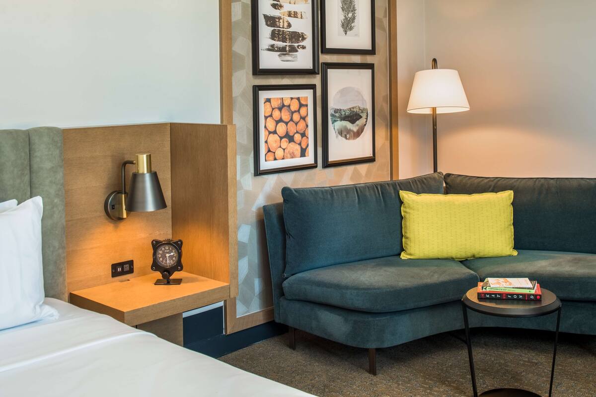 A hotel room with a bed, a nightstand with a lamp, a sofa with cushions, a small side table with books, and framed pictures on the wall.