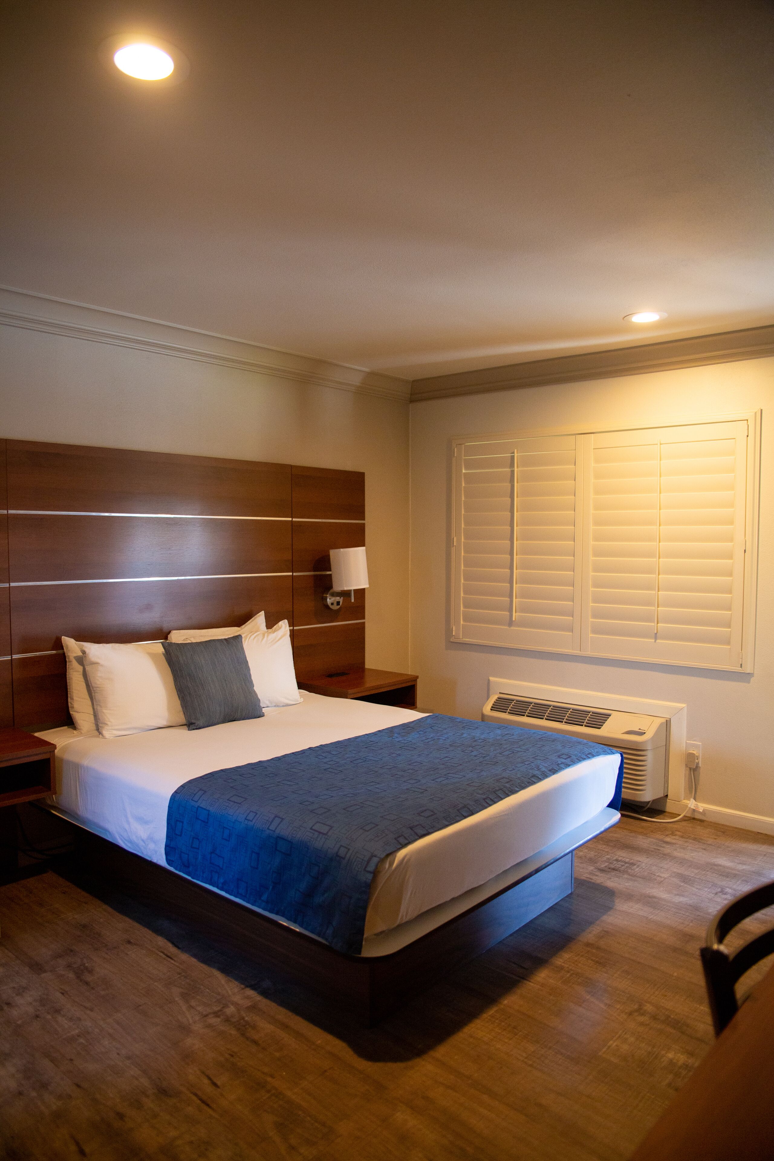 A bedroom with a wooden headboard, a bed with a blue blanket, a bedside lamp, and a window with blinds.