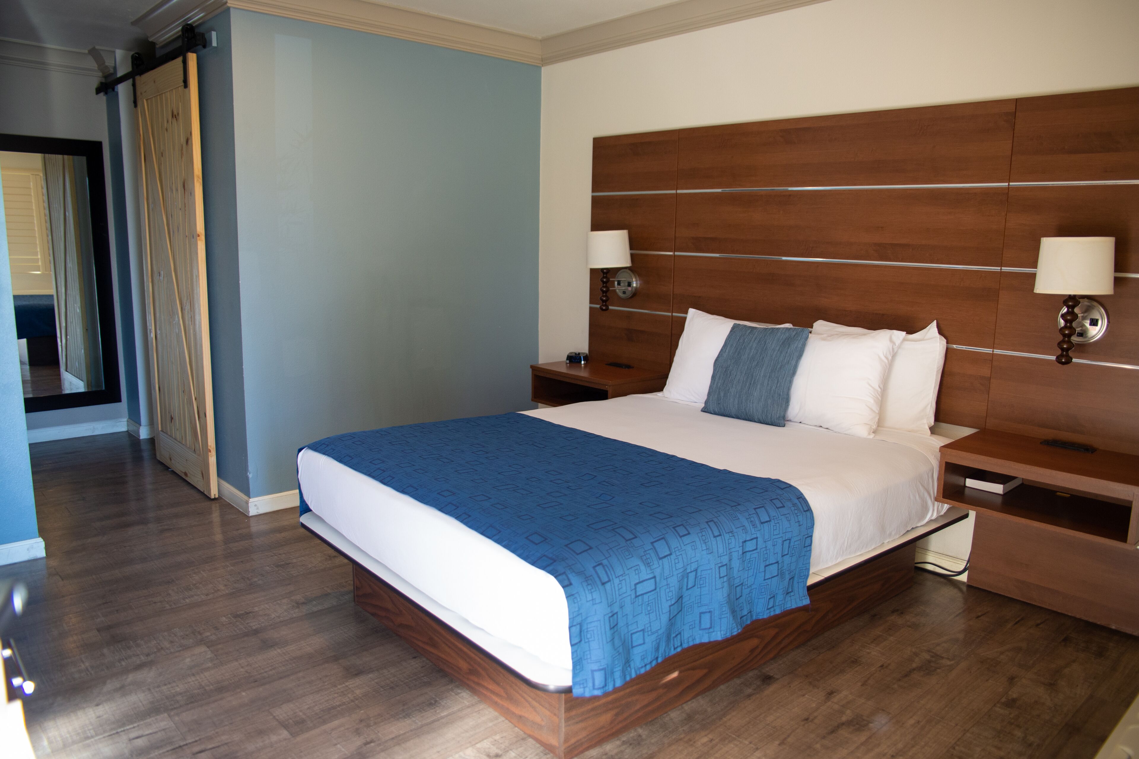 A hotel room with a large bed, wooden headboard, two bedside tables with lamps, and a blue bedspread.