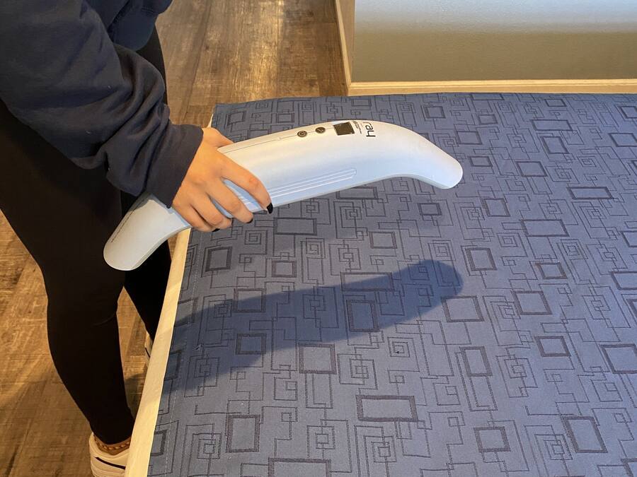 Una persona sosteniendo un objeto blanco, posiblemente una herramienta de limpieza, en una habitación con alfombra estampada y suelo de madera.