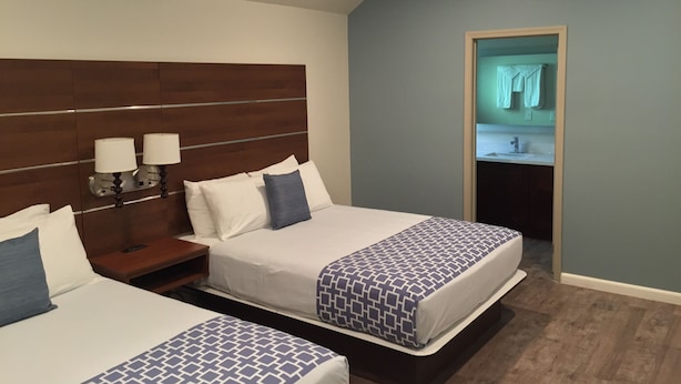 A hotel room with two beds, a wooden headboard, bedside lamps, and a view of a bathroom through an open door.