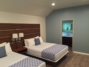 A hotel room with two beds, a wooden headboard, bedside lamps, and a view of a bathroom through an open door. - Sterling Inn (Santa Clara)