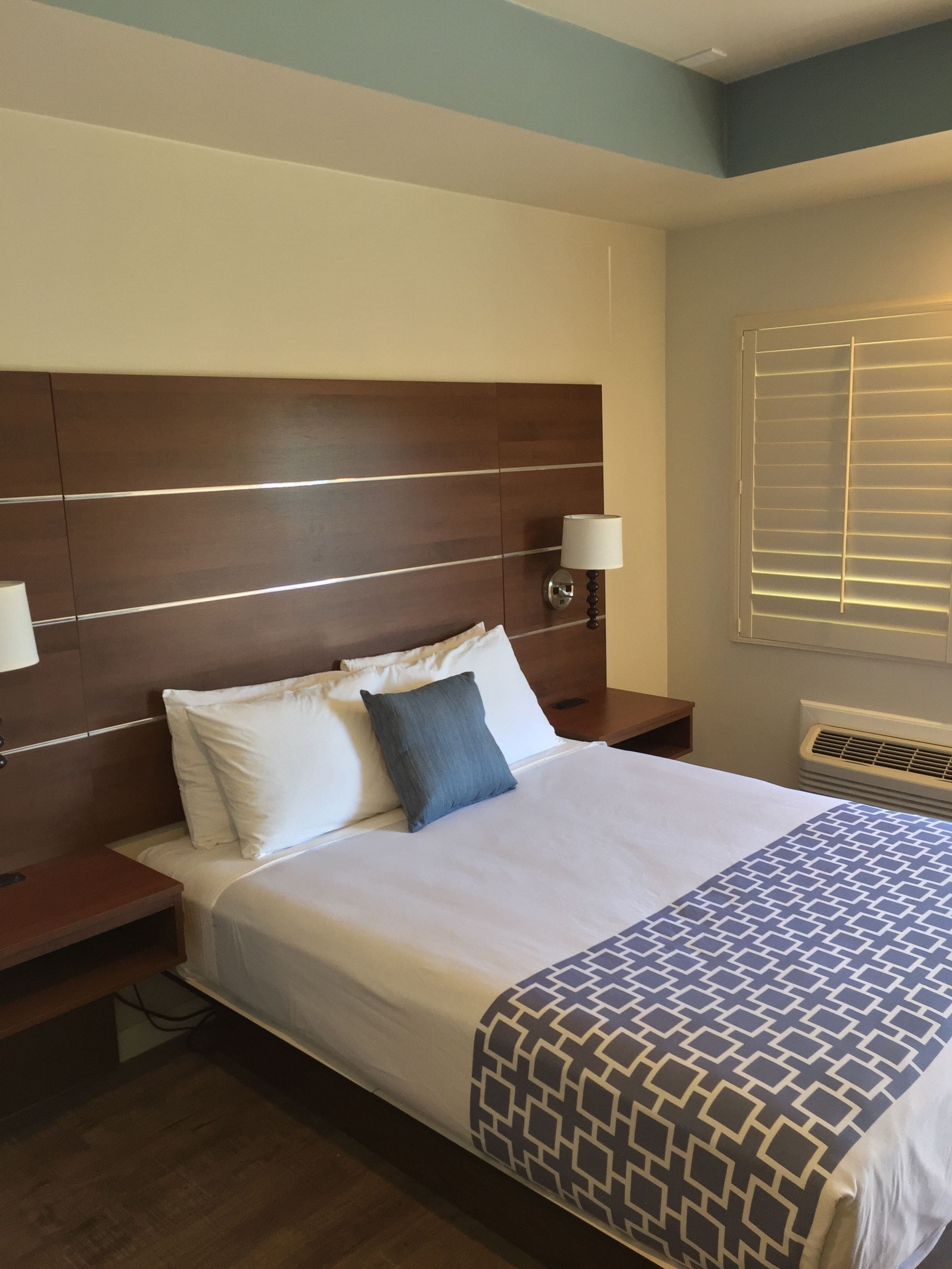 A hotel room with a large bed, a wooden headboard, bedside lamps, and a window with blinds.