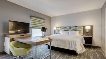 A modern hotel room with a bed, desk, chair, TV, and artwork on the wall.