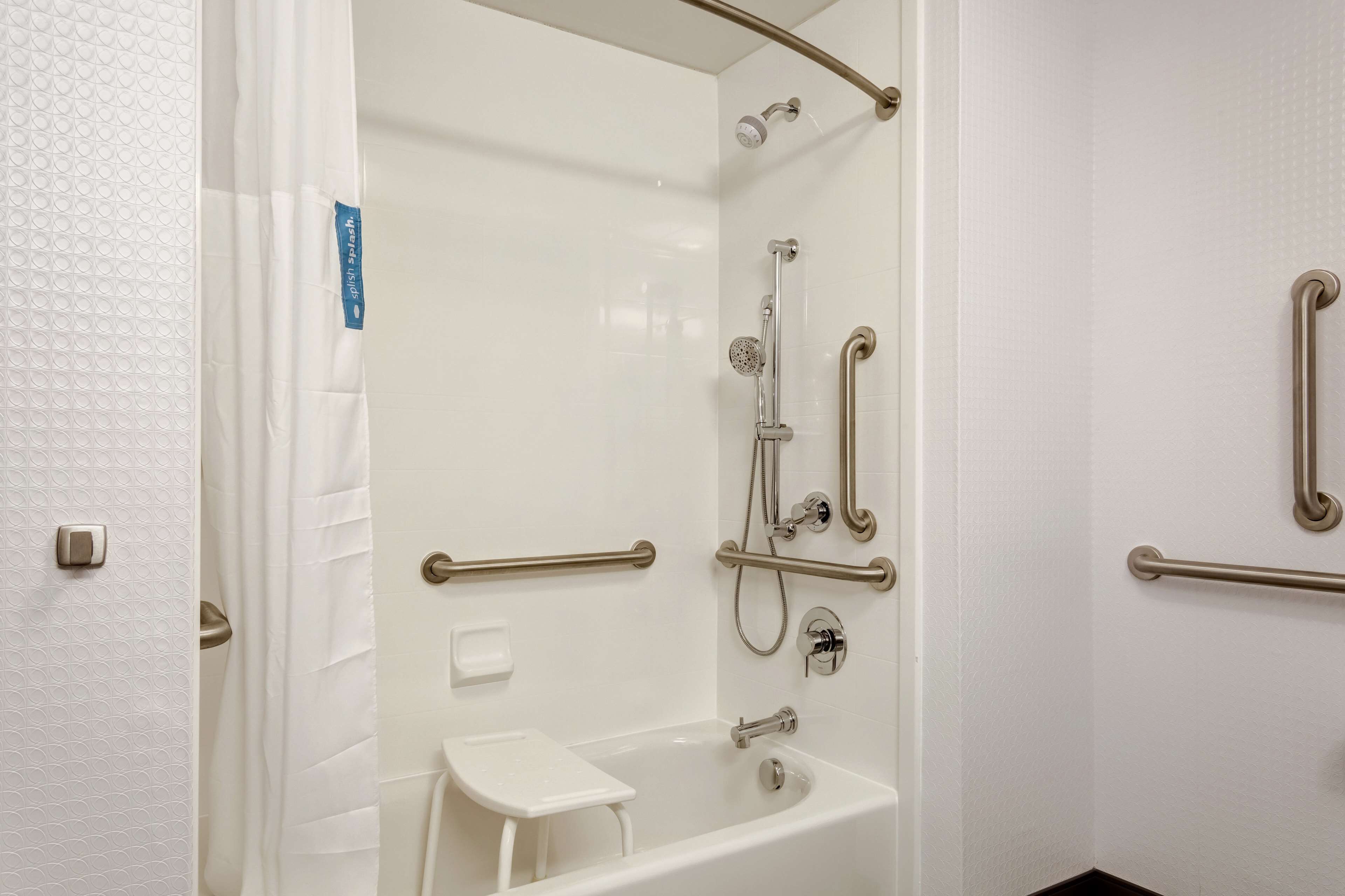 Une salle de bain avec une douche à l’italienne, des barres d’appui et un petit tabouret blanc.