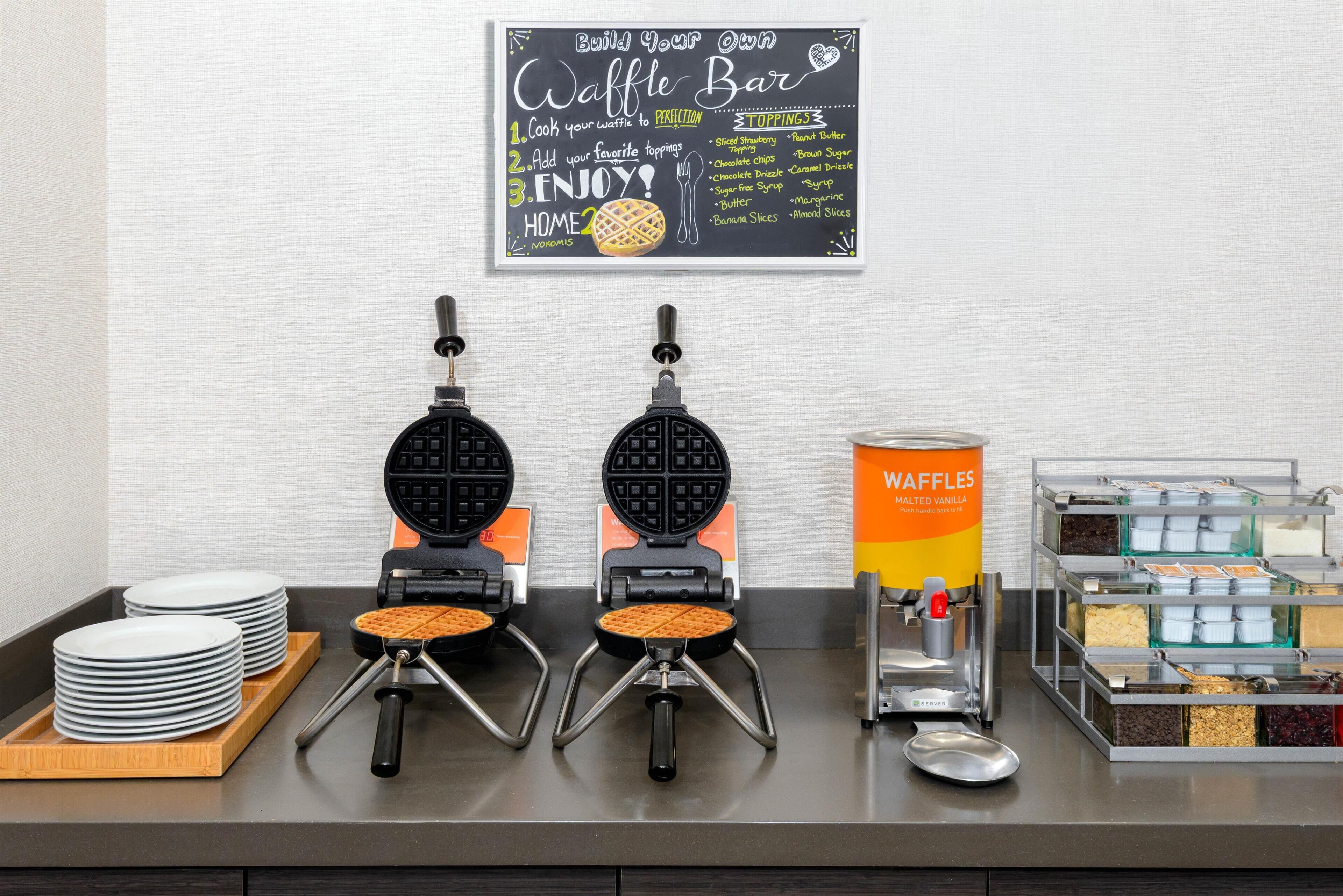 A waffle station with two waffle makers, a waffle container, and a sign listing waffle toppings.