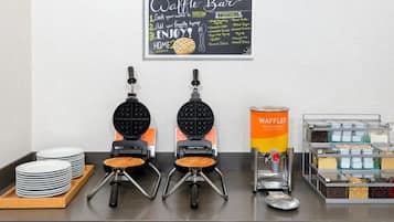 A waffle station with two waffle makers, a waffle container, and a sign listing waffle toppings.