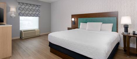 Habitación de hotel con una cama grande, una mesita de noche con lámpara, un televisor sobre un mueble bajo y una ventana con cortina estampada.