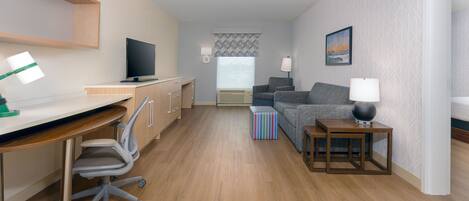 A modern hotel room with a wooden desk, a television, a sofa, a patterned ottoman, and a bedside table with a lamp.