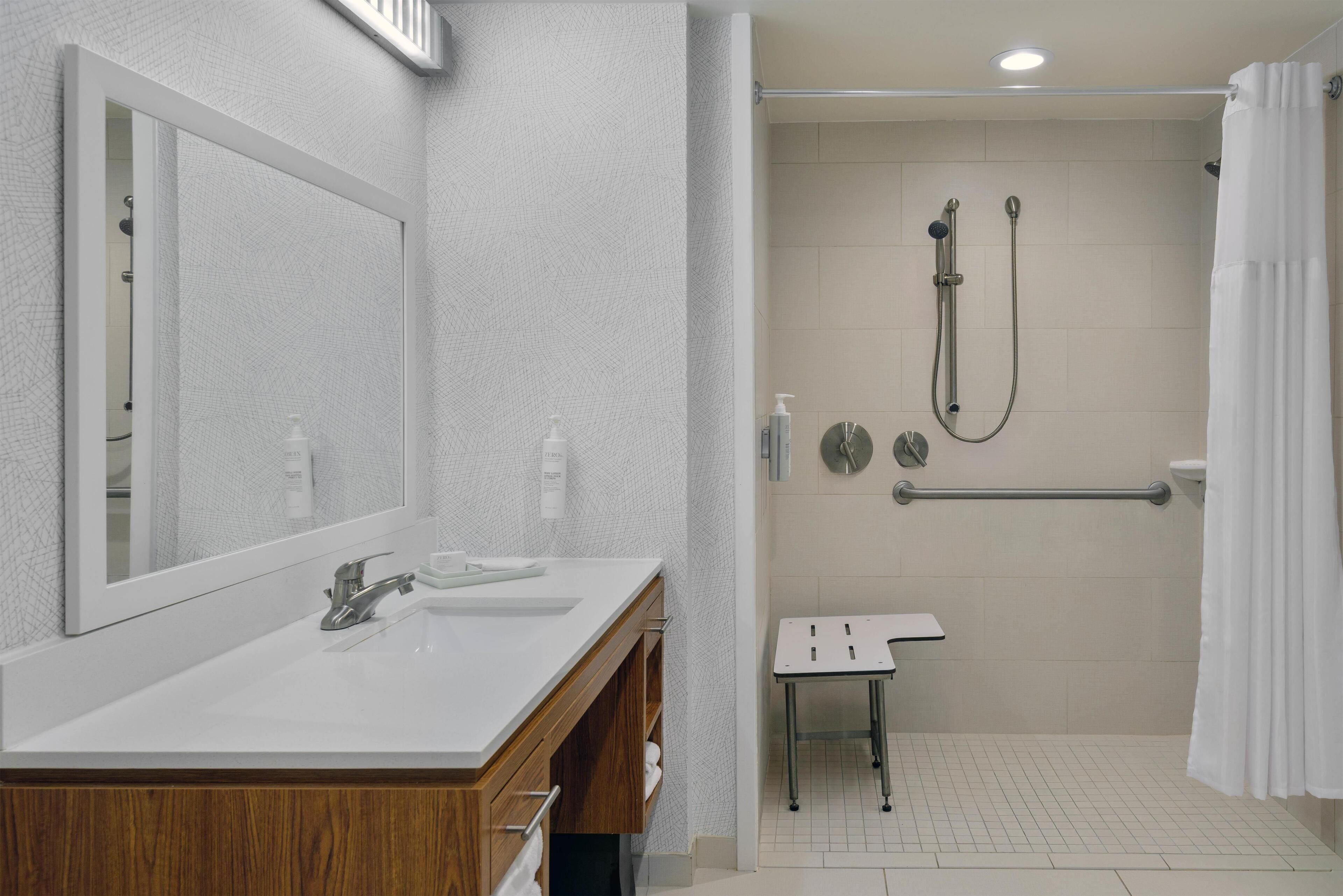 A modern bathroom with a large mirror, dual sinks, a shower with a handheld showerhead, and a grab bar.
