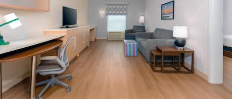 A modern hotel room with a wooden desk, a television, a sofa, and a small table with a lamp.