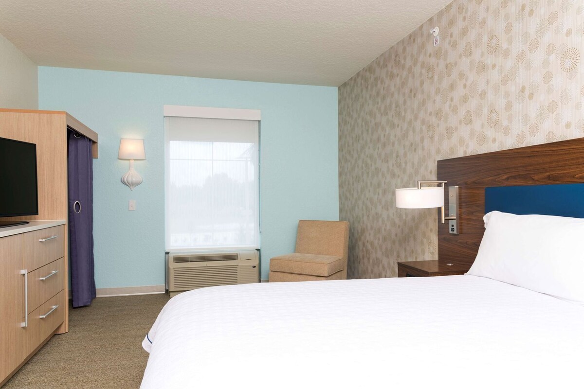 Habitación de hotel con cama, televisión, una silla, ventana con persianas y una lámpara fijada a la pared.