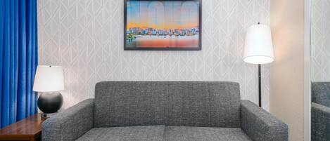 A grey sofa with a patterned cushion, a framed cityscape picture on the wall, a lamp on a side table, and a blue curtain.